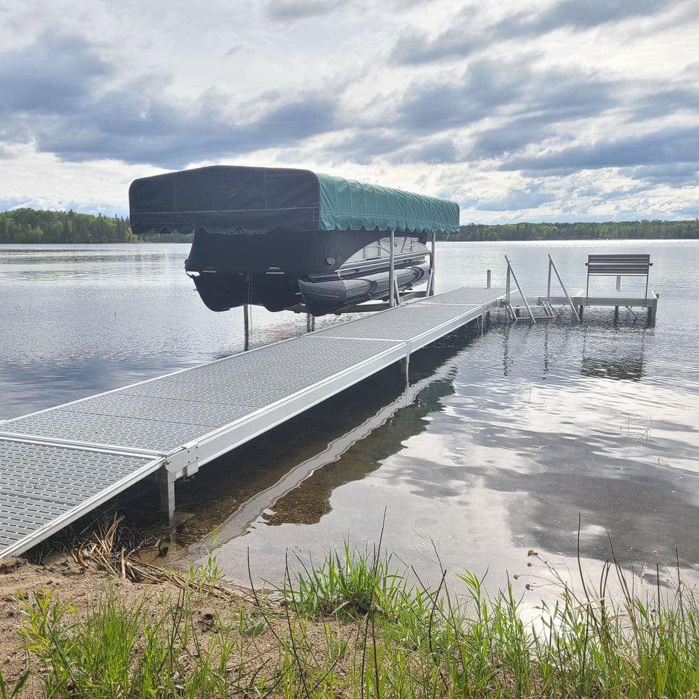 Pontoon boat on a dock by a tranquil lake with cloudy skies and lush greenery.